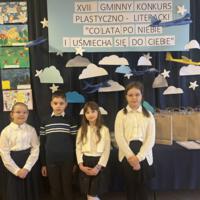 Dzieci oglądające prace. W tle na granatowej kotarze chmury oraz samoloty