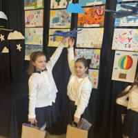 Dzieci oglądające prace. W tle na granatowej kotarze chmury oraz samoloty